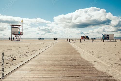 Fototapeta Naklejka Na Ścianę i Meble -  Wide, sandy beach with a wooden walkway leading to the shore. There are lifeguard stations and people walking along the beach. The sky is partly cloudy with scattered clouds