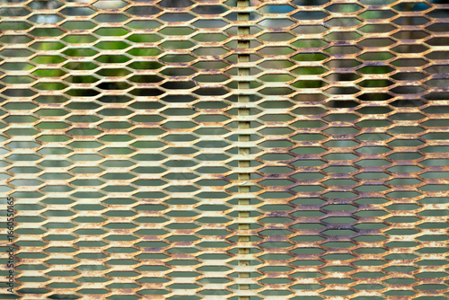 A metallic mesh screen with blurred reflections of greenery behind it. A textured metal fence with light and shadow effects.