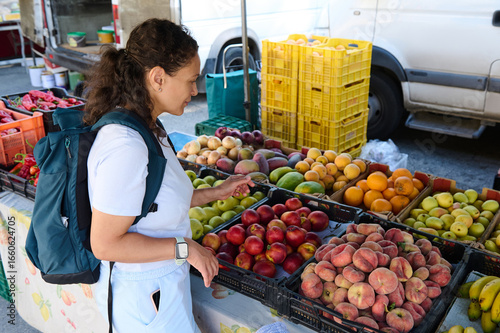 A woman with a backpack examines fresh fruits and vegetables at an outdoor local market. The scene reflects healthy lifestyle choices and the vibrant colors of fresh produce in natural settings.