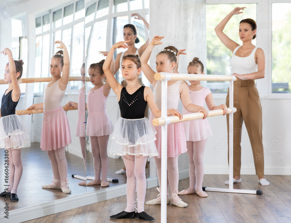 Obraz premium Adult female teacher corrects group of girls dancers standing in first position at ballet barre