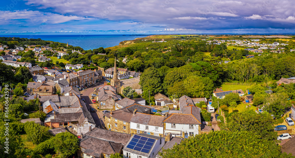 Fototapeta premium Aerial view of St Agnes village in Cornwall, England, with stone houses, church spire, lush greenery, and coastline in the background