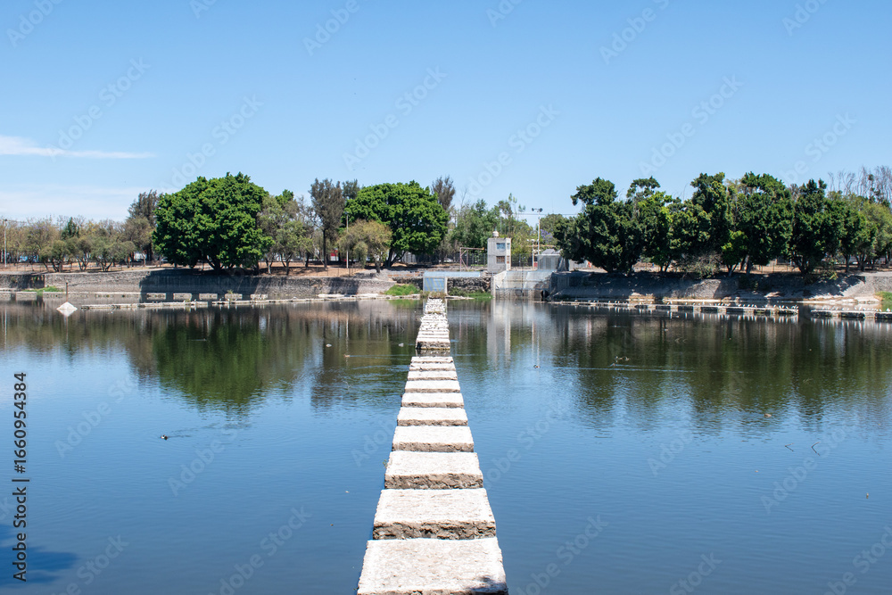 Fototapeta premium Long stone path stretches across a calm body of water at Parque Independencia El Dean in Guadalajara, Mexico under a clear sky