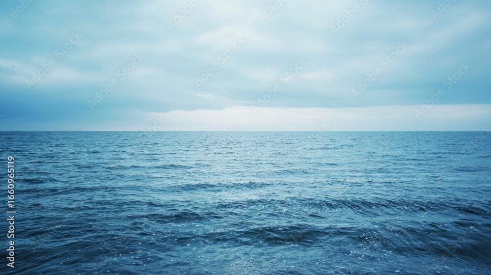 Obraz premium A wide, expansive view of a dark blue ocean meeting a cloudy, pale blue sky. Gentle waves ripple across the water's surface, reflecting the muted light.