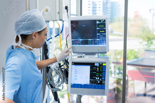 Nurse monitoring patient vital signs on dual screens beside large window in hospital room