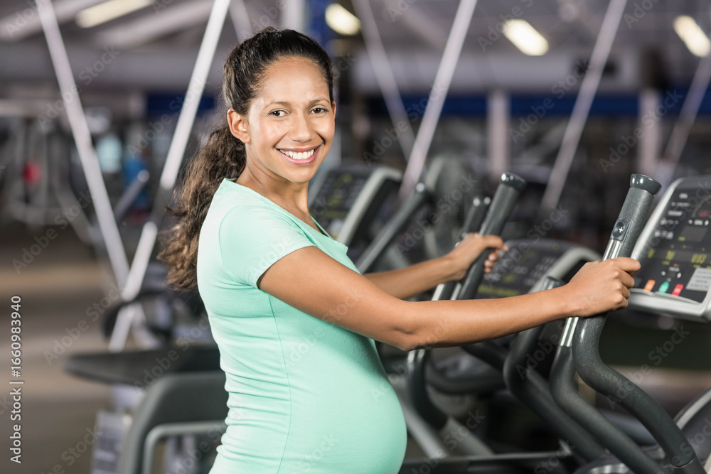 Fototapeta premium Pregnant woman standing on elliptical machine in gym using console and cardio equipment
