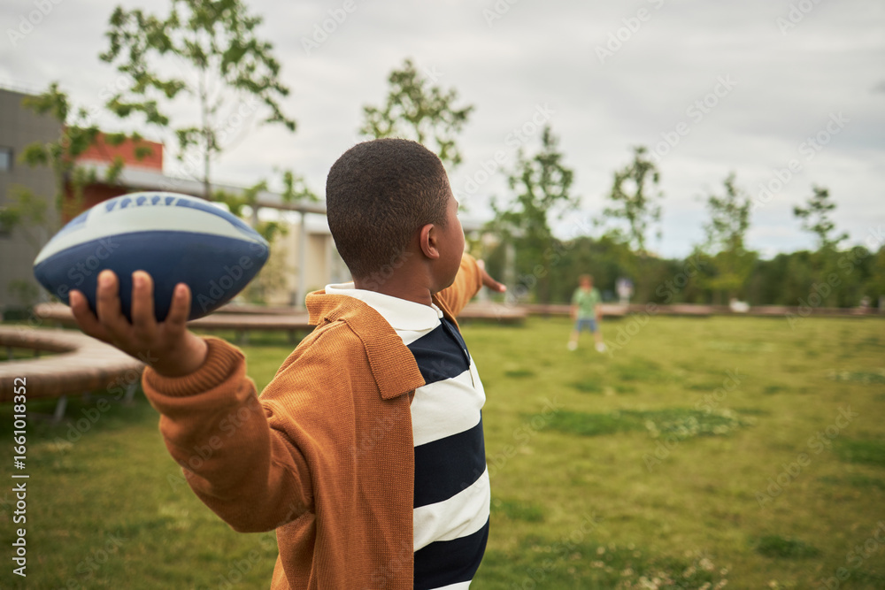 Fototapeta premium Black boy throwing football outdoors on grassy field, extending arm back in preparation to pass, another child visible in distance, casual playtime activity in park setting