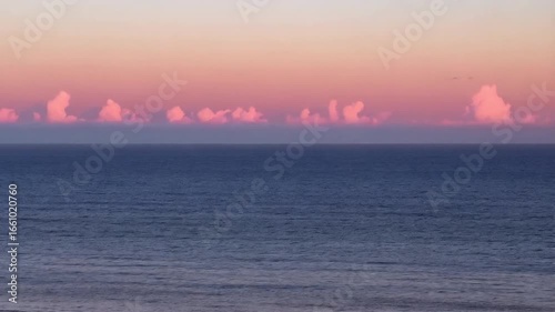 Gentle pastel clouds drift across the horizon above a calm, deep blue ocean at dawn.