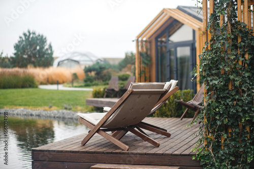Two modern, comfortable lounge chairs on a wooden deck next to a pond, with a blurred backdrop of a contemporary vacation house and natural landscape.
