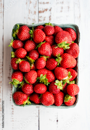 Carton of fresh strawberries on white wood backdrop from above.