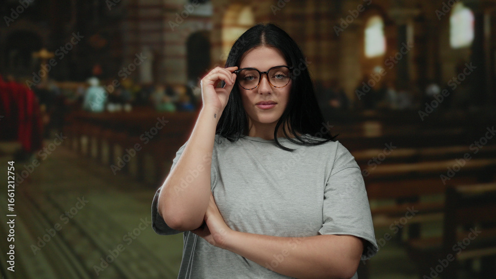 Fototapeta premium Young woman with glasses standing confidently in a church, wearing casual attire and expressing empowerment through her pose in an indoor religious setting.