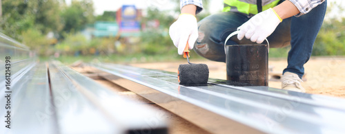 Fotografie Construction worker painting steel beam with black protective coating using roller brush