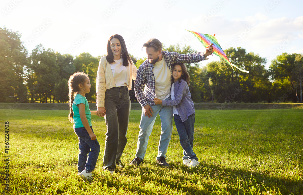 Fototapeta premium Summer green park, happy family standing smiling warmly, holding preparing colorful kite for flying, feeling joyful and connected embracing on sunny weekend day, playing game together for fun