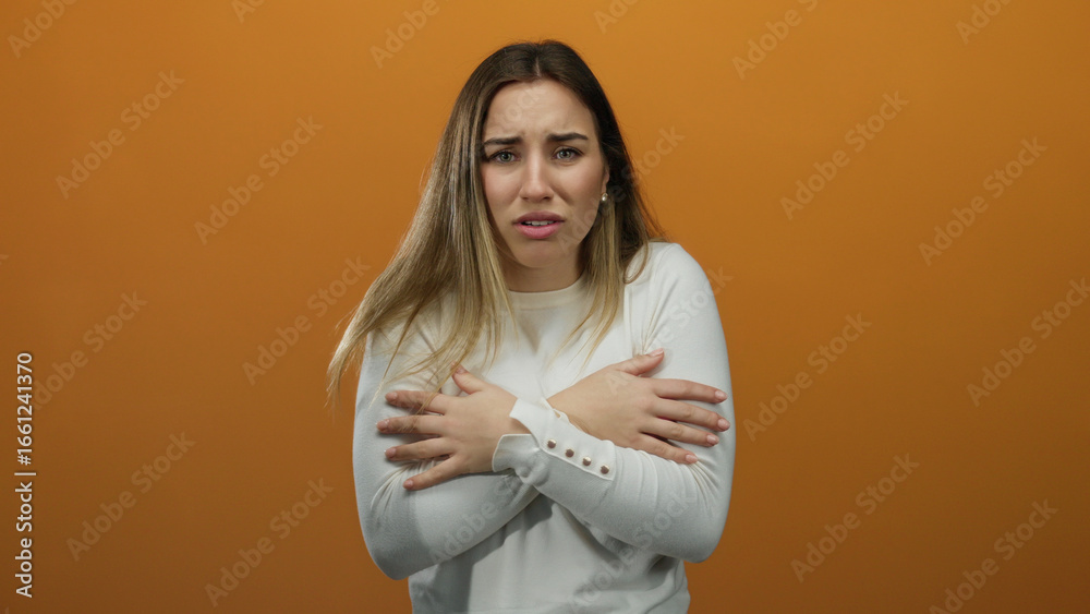 Obraz premium Young blonde woman feeling cold in a white sweater, hugging herself against an isolated orange background, expressing discomfort and shivering slightly.