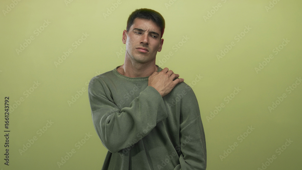 Obraz premium Young hispanic man grimacing with hand on shoulder against green studio wall wearing olive sweater; pain.