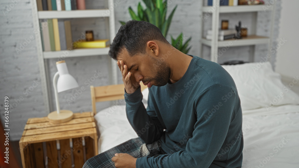 Obraz premium Young hispanic man sitting on bed with hand on forehead in cozy bedroom, displaying stress or fatigue, surrounded by books and plants suggesting a home interior setting.