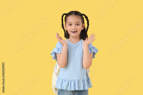 Cute little shocked Asian girl with backpack on yellow background. End of sch...