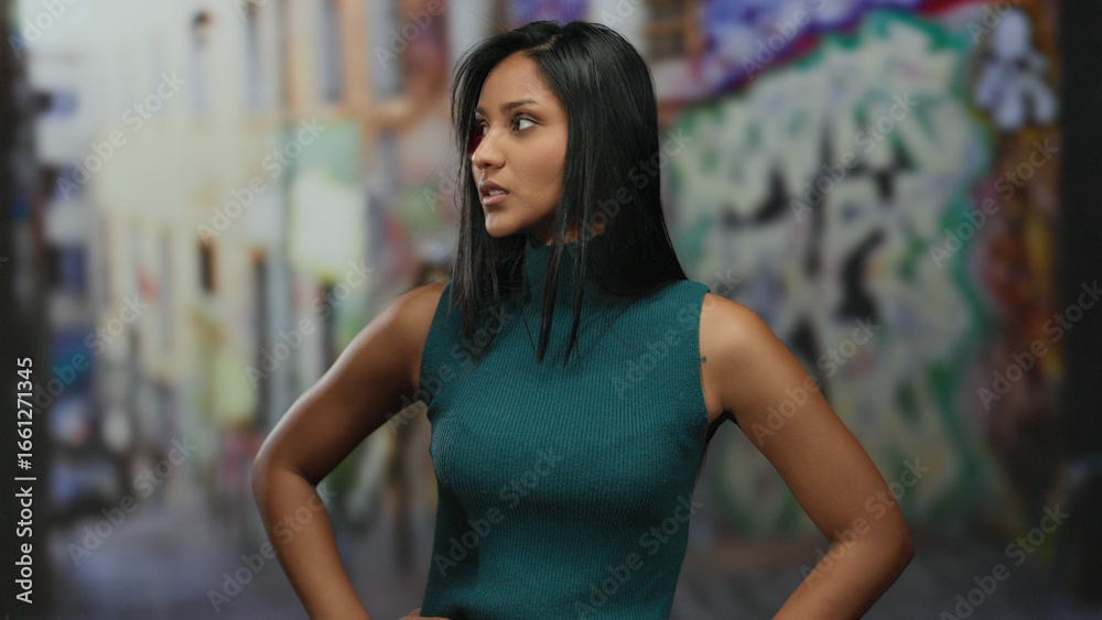 Fototapeta premium Woman standing outdoors in urban street with graffiti background, expressing emotions with hands, dressed in teal, latina identity, candid mood, cityscape vibe.