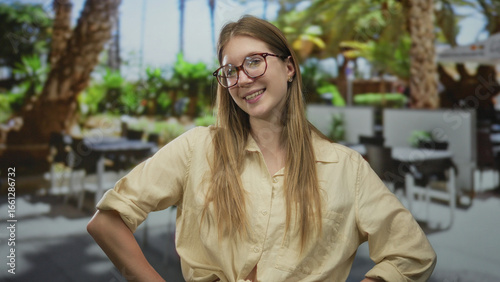 Woman stands with hands on hips on street in light beige shirt and round glasses under sunlight with blurred palm trees; confidence.