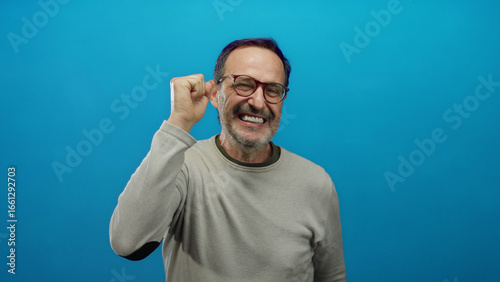 Mature hispanic man expressing joy and excitement with raised hands against a vibrant blue background, showcasing an enthusiastic and animated expression.