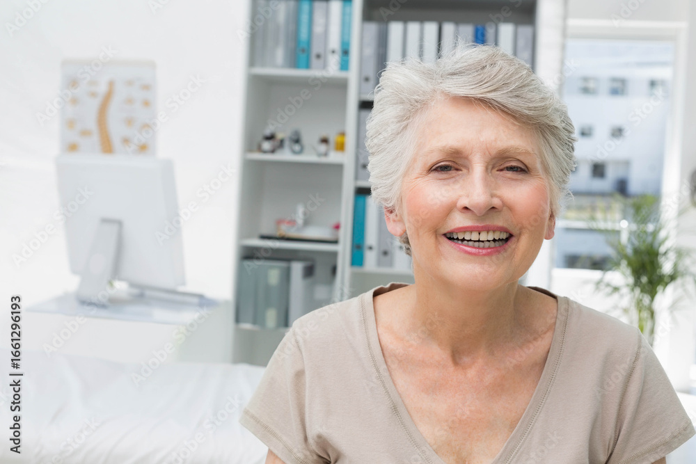 Obraz premium Senior female sitting and smiling in clinic office with monitor, spine chart, plant, copy space