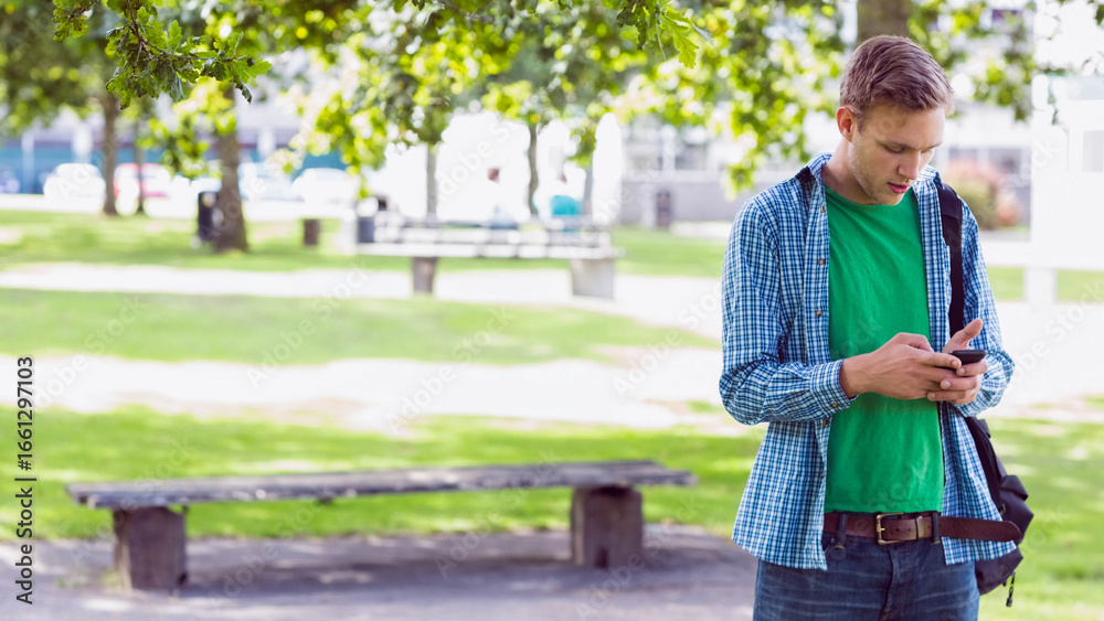 Fototapeta premium Man standing under leafy tree shade in urban park, checking smartphone beside wooden bench