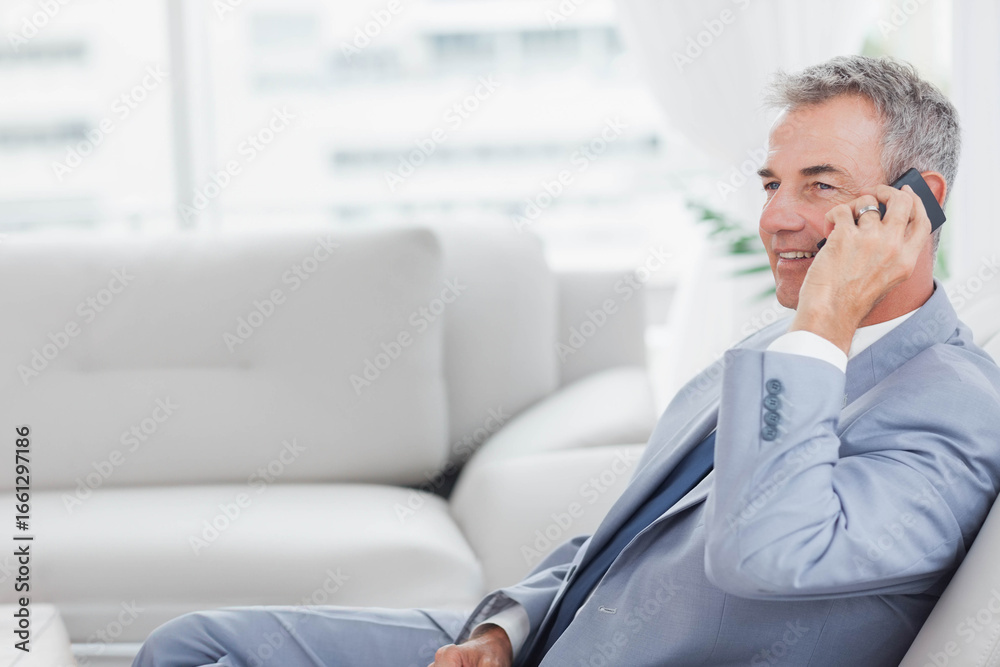 Fototapeta premium Senior man in suit sitting on sofa by plant talking on smartphone at reception, copy space
