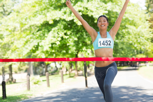Asian woman running through red finish line tape on park path with turquoise top bib 1452