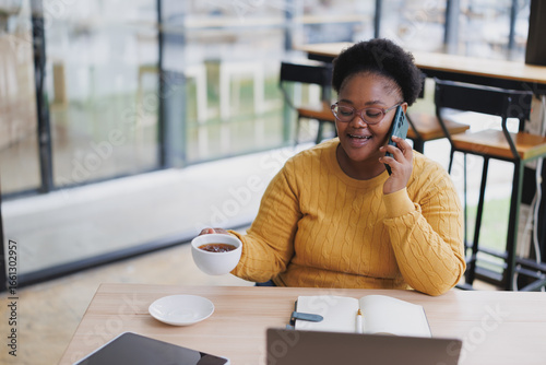 Cheerful african american businesswoman is working remotely in a cafe, talking on the phone and drinking tea, with a laptop and tablet on the table
