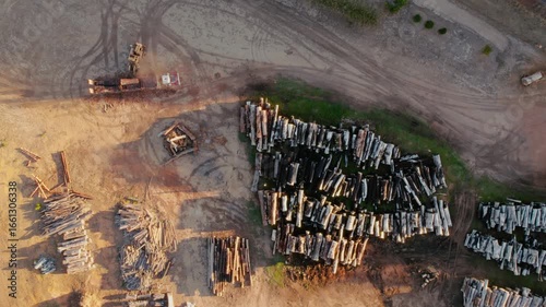 Aerial view of a heavy fork lifter offloading a truck with logs at lumber yard filled with stacked timber, and heavy machinery on a sunny afternoon