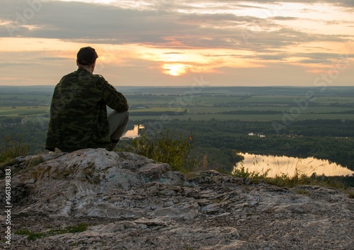 The silhouette of a man in camouflage sitting on a mountaintop, with a river and a sunset in the background