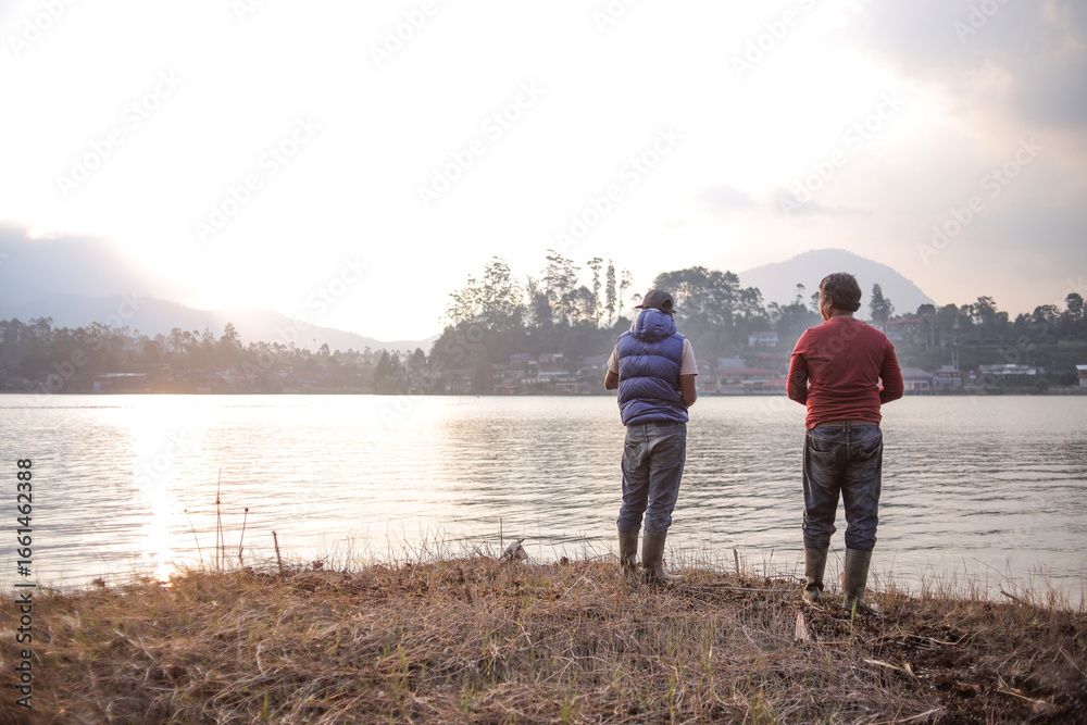 Fototapeta premium Two Men Standing on a Grassy Bank at Sunrise Fishing in a Lake With a Scenic View of Mountains