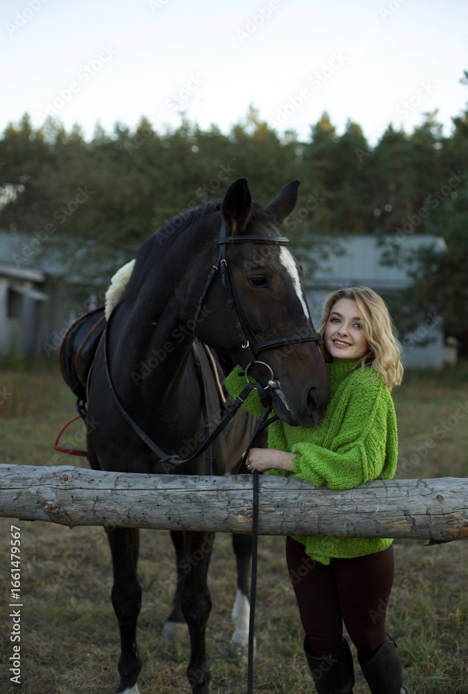 Obraz premium A woman in a green sweater is smiling while standing beside a black horse. They are positioned near a wooden fence on a peaceful rural setting during the late afternoon.