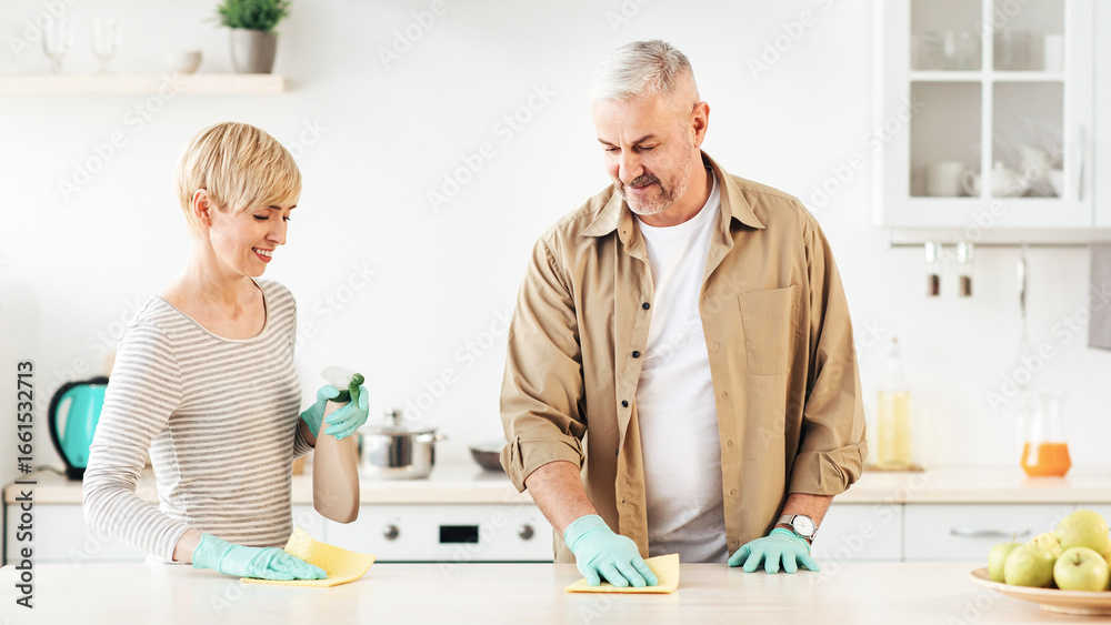 Obraz premium Smiling woman and man with detergent spray wiping dust off from kitchen table with rag. Adult family couple in blue gloves cleaning up in modern white cozy apartment interior together, copy space