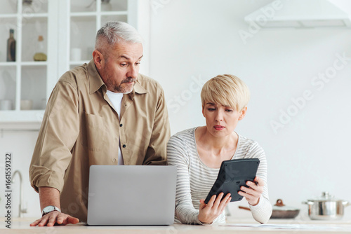 Family economics, bookkeeping, bill payment and taxes. Surprised upset adult wife shows calculator to husband, man pays bill on laptop in kitchen interior, empty space