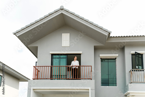 Modern two-story house with balcony and gabled roof. Man stands at railing by sliding windows. Contemporary home facade shows residential architecture, exterior design, realty, and property style.