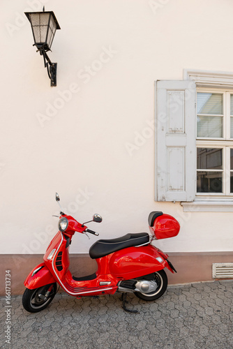 Red scooter parked by a white wall with window and lantern, urban street scene