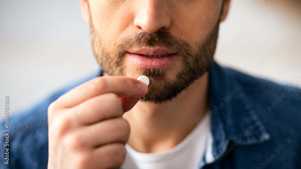 Fototapeta premium Treatment, medicine, medication, pills, vitamins, supplements concept. Cropped of bearded man taking white pill. Closeup of unrecognizable man holding medicine next to his mouth