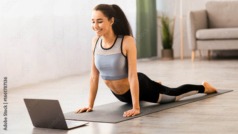 Fototapeta premium Fitness online. Athletic young woman doing yoga in living room, stretching on yoga mat in front of laptop, copy space. Good looking brunette lady exercising at home, using video lessons, side view