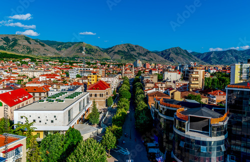 Vibrant Panoramic View of Korçë, Albania with Colorful Rooftops, Tree-Lined Boulevards, and Majestic Mountain Range Under a Clear Blue Summer Sky