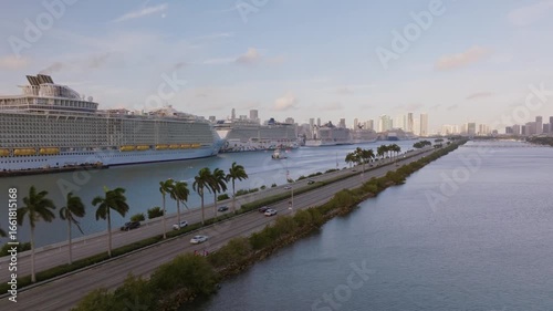 Aerial reveal shot of MacArthur Causeway with cars driving near Miami Port, luxury ships docked, and Miami downtown skyline at sunset over Biscayne Bay