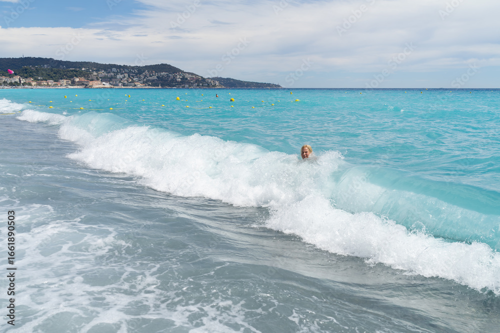 Fototapeta premium A happy, joyful blonde woman plays and swims in the beautiful turquoise waves of the Mediterranean Sea in Nice, France.