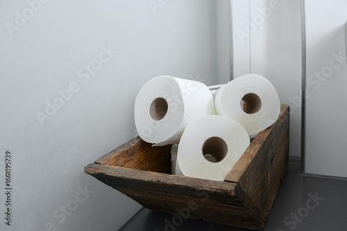 neatly arranged rolls of white toilet paper stored in a rustic wooden basket placed against a minimalistic gray wall, highlighting practicality and design in bathroom decor