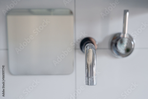 top-down view of a sleek chrome bathroom faucet beside a circular handle and a square mirror mounted on a white tiled wall, highlighting contemporary design and minimalism