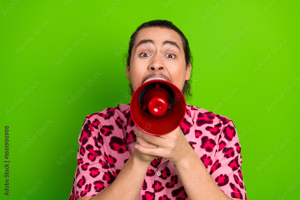 Fototapeta premium Young man with red megaphone in pink leopard-print shirt exclaiming vividly against green background during a vibrant moment