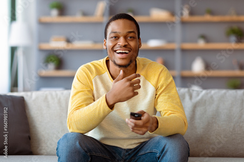 Foto Laughing black guy watching comedy on TV at home, sitting on sofa in living room, holding TV remote