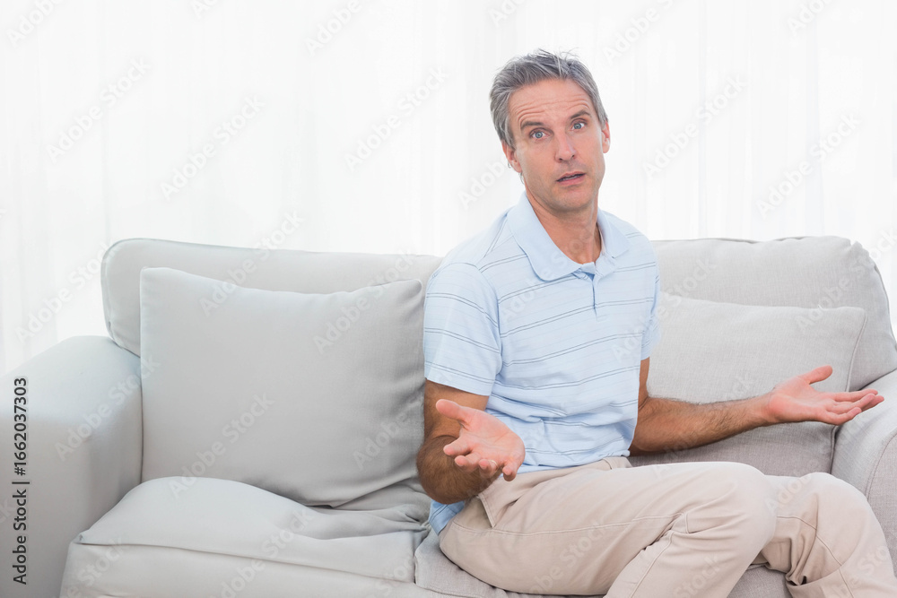 Man raising palms asking question seated on sofa in living room with cushions, curtains, copy space Man raising palms asking question seated on sofa in living room with cushions, curtains, copy space