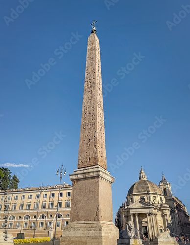View of the towering Egyptian obelisk in Rome, Piazza del Popolo. The image highlights its ancient hieroglyphs and a classical domed church, all set against a clear blue sky