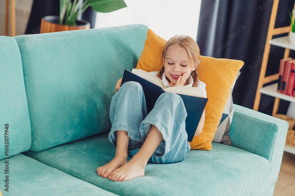 Obraz premium Happy young girl reading a book on a colorful couch in a cozy living room setting, enjoying a peaceful leisure afternoon.