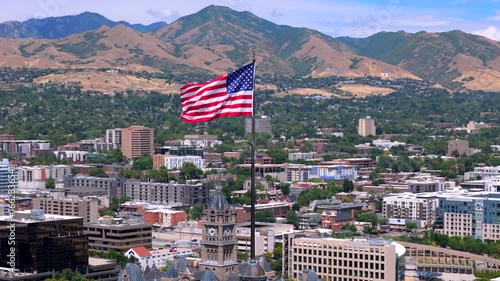 Aerial footage shows an American flag waving on a tall pole, with a vibrant Salt Lake City, Utah and majestic mountains in the background, symbolizing freedom and patriotism for all viewers