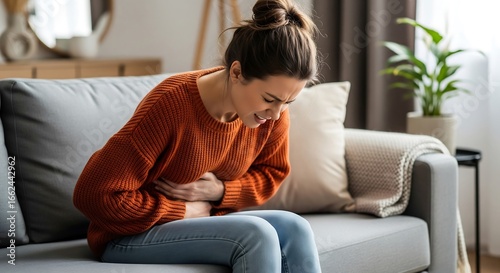 A young woman sits on a grey couch, clutching her stomach in apparent pain. She is wearing an orange sweater and jeans, exhibiting signs of discomfort or illness. Her expression is one of di